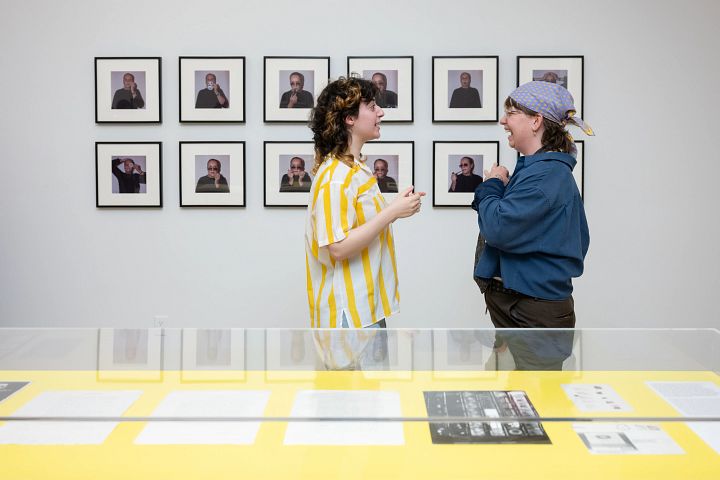 Dos personas conversan de pie dentro de El Museo, situadas detrás de una vitrina amarilla y frente a una cuadrícula de pequeños retratos enmarcados.