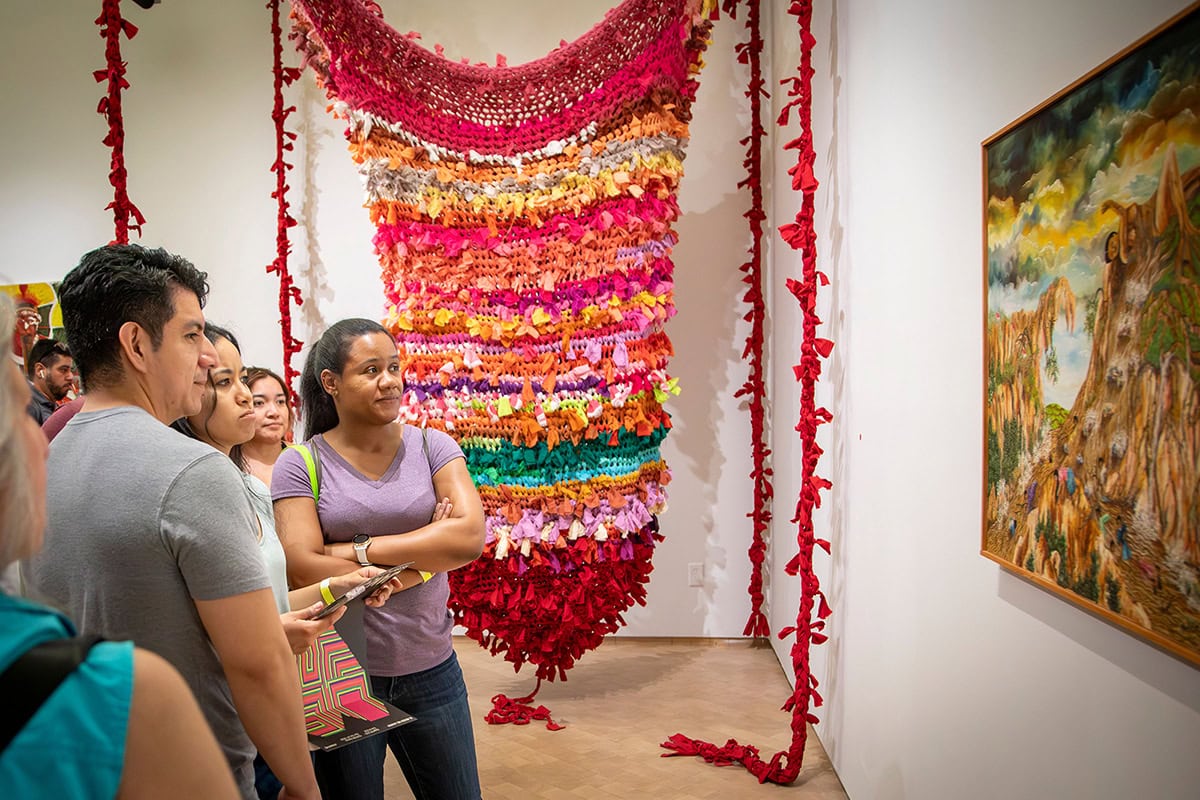 Un grupo de personas permanece de pie dentro de una galería de arte, mirando atentamente una pintura en la pared junto a una gran y colorida instalación de tejido textil.