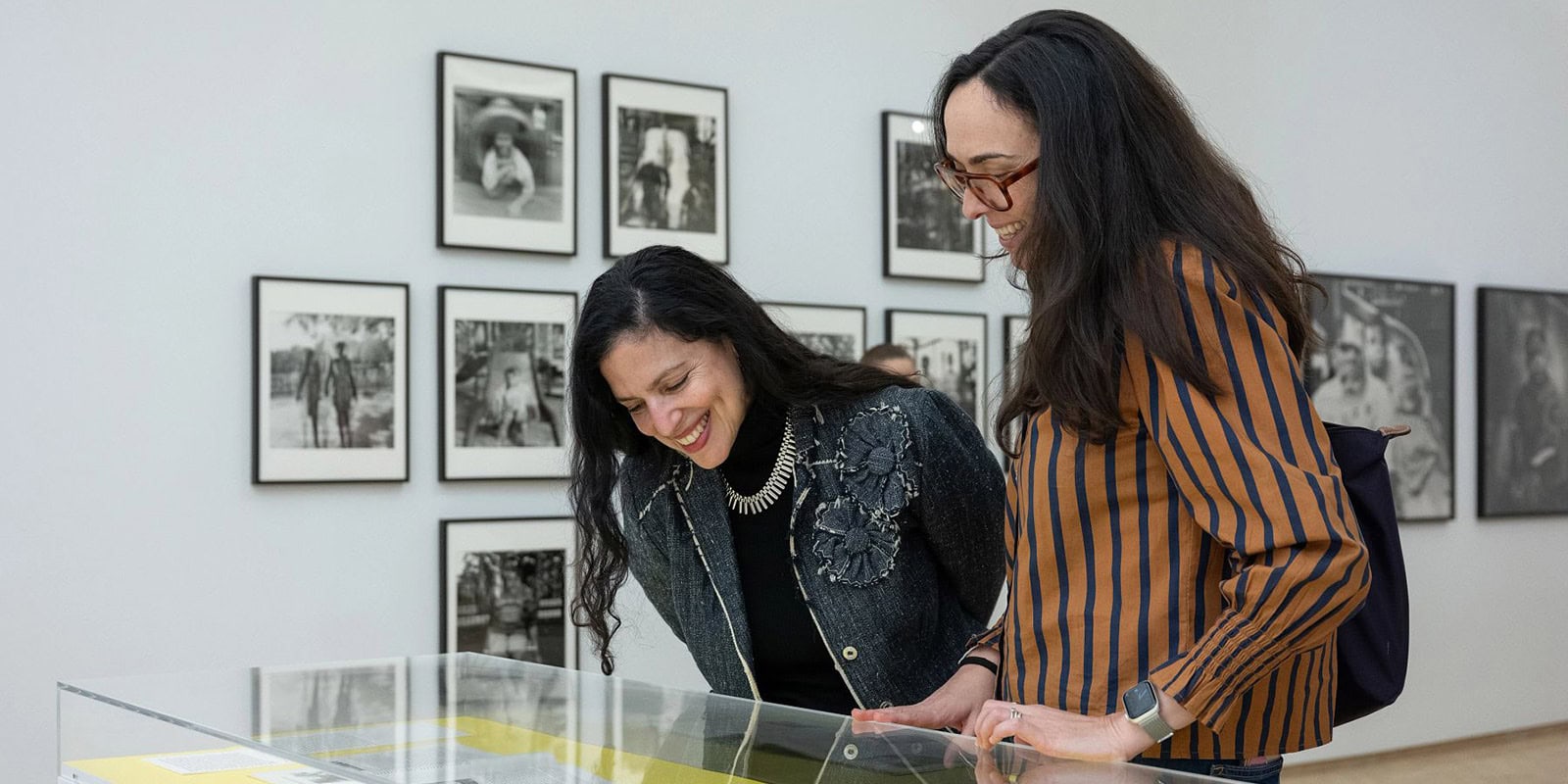 Dos mujeres sonrientes miran hacia abajo a una vitrina dentro de El Museo con fotografías en blanco y negro enmarcadas colgadas en la pared detrás de ellas.