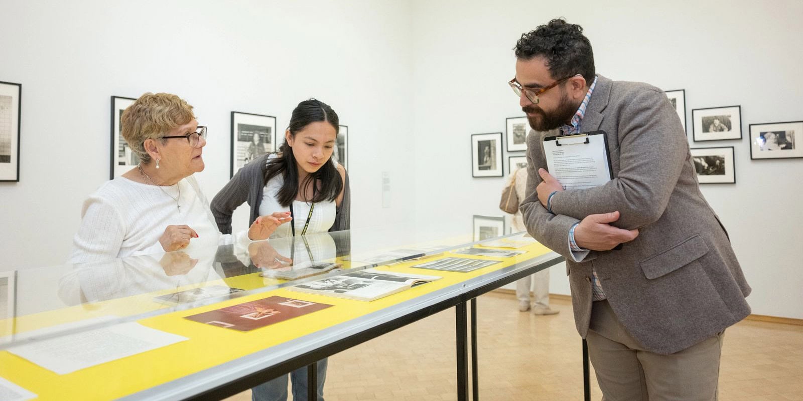 Two women look at documents inside a long display case while a man holding a clipboard listens to them inside El Museo.