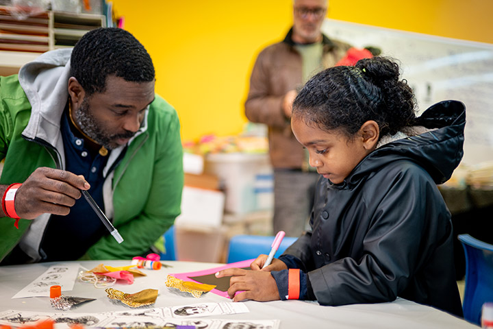 A man leans in to watch a young girl draw on a piece of paper with a marker surrounded by art supplies, while another man stands blurred in the background.