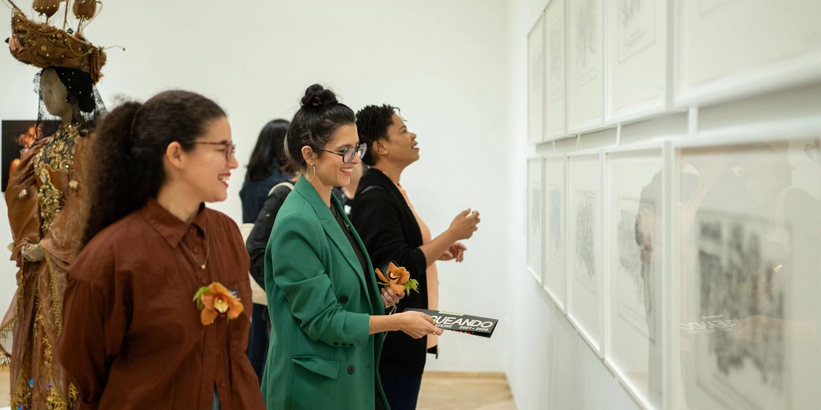 A group of women stand inside El Museo, smiling as they look at a series of framed artwork hanging on a white wall.
