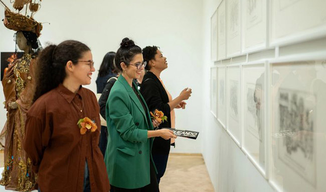 A group of women stand inside El Museo, smiling as they look at a series of framed artwork hanging on a white wall.