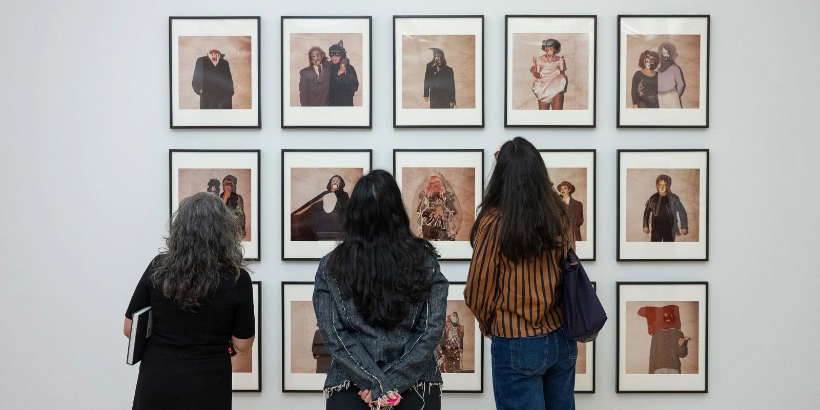 Tres mujeres están de espaldas a la cámara, mirando filas de retratos enmarcados de figuras enmascaradas y disfrazadas colgados en una pared blanca de la galería.