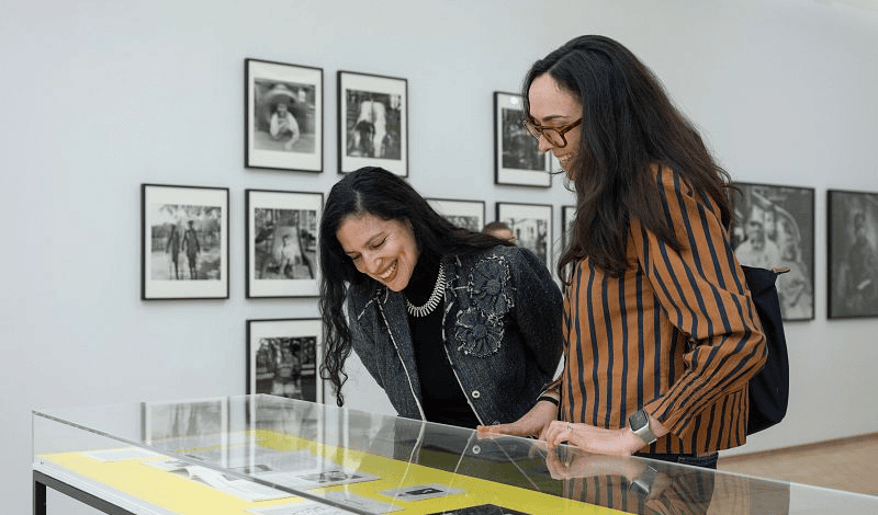 Two smiling women look down at a display case inside El Museo with framed black-and-white photographs hanging on the wall behind them.