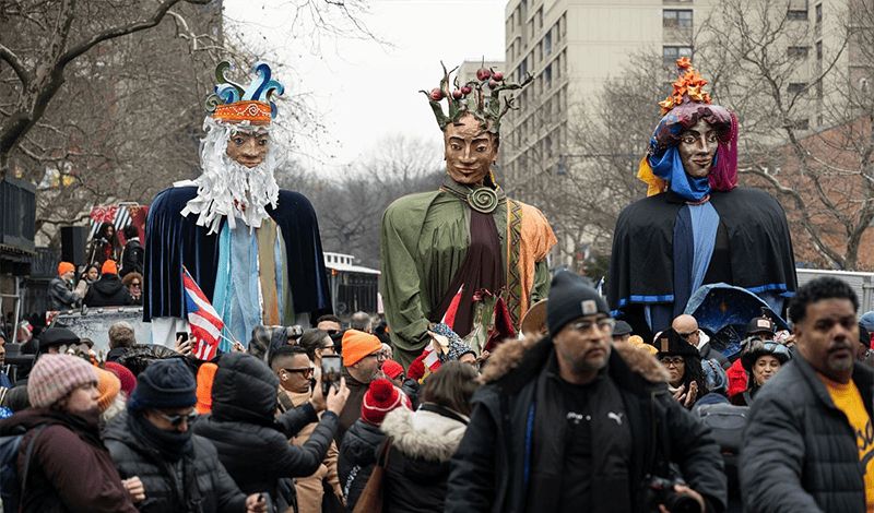 Tres figuras de marionetas gigantes que representan a los Reyes Magos se elevan sobre una concurrida calle de la ciudad durante el desfile festivo.