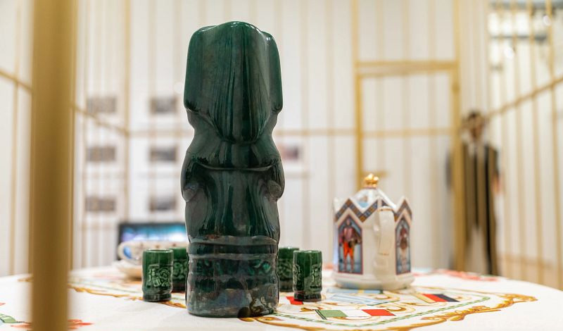 A green sculpture and a decorative ceramic pitcher sit on a table inside El Museo display space with golden bars and blurred background figures.