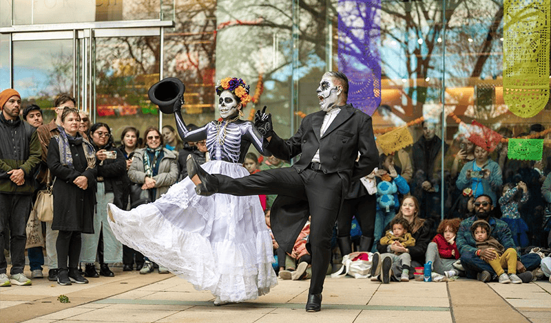 Two performers dressed and painted as skeletons dance dynamically in front of an outdoor crowd gathered to watch them.