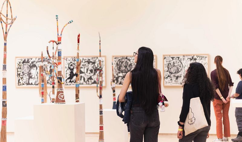 Two women stand in an art gallery looking at tall, slender sculptures on pedestals and framed black-and-white artworks hanging on the wall.