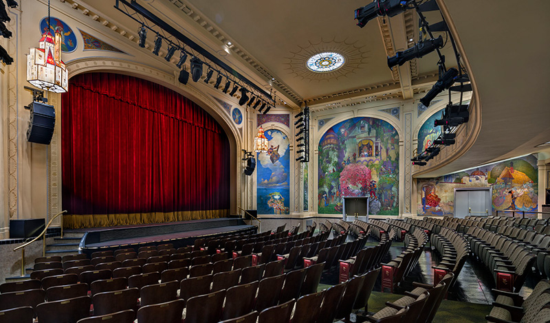 La fotografía muestra el interior de un teatro decorado de forma vistosa, con filas de asientos vacíos frente a un escenario con un gran telón de terciopelo rojo, rodeado de coloridos murales en las paredes.