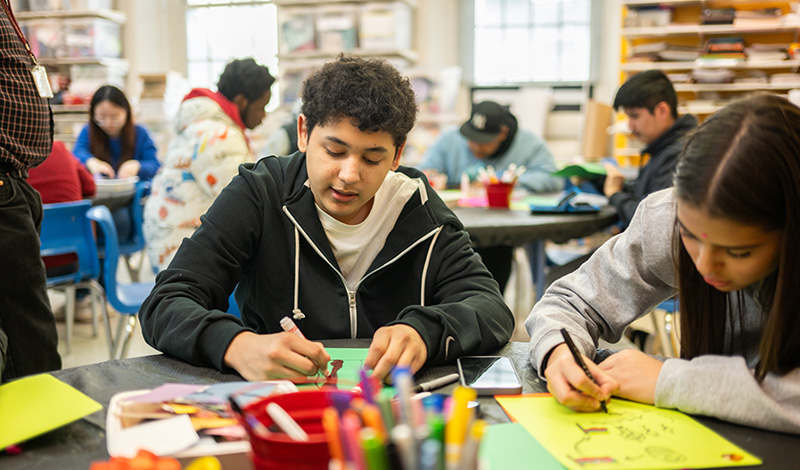 A group of students work on art projects at tables filled with colorful paper and markers inside a brightly lit classroom.