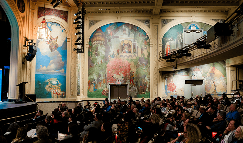 An audience sits in an ornate theatre watching a presentation on a stage next to walls decorated with large, colorful murals.