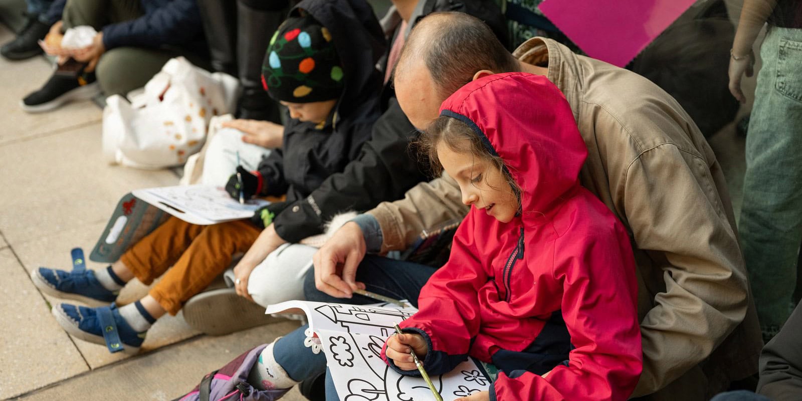 A man and a young girl sit together outdoors coloring in a coloring book alongside other children sitting on the pavement.