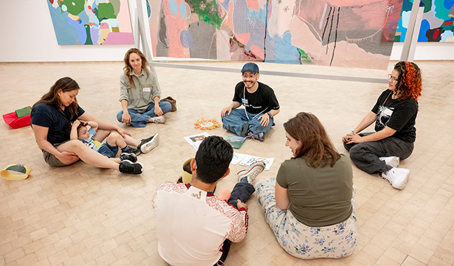 Un grupo de personas, incluido un niño pequeño, se sientan con las piernas cruzadas en un círculo sobre el suelo de El Museo rodeados de pinturas grandes y coloridas.