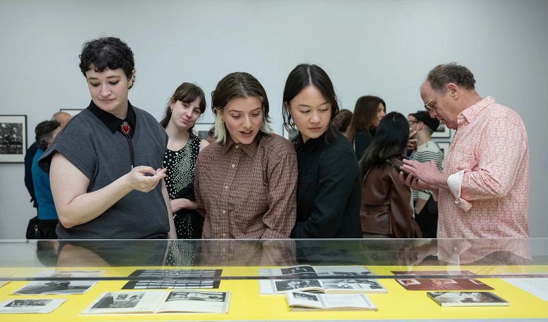 A group of people look down at open books and documents displayed inside a glass case with a yellow background at an art exhibition.