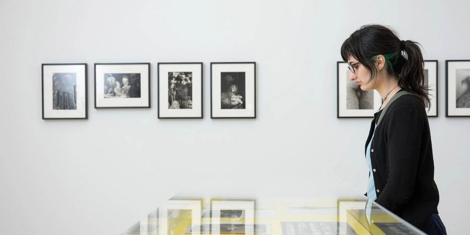 Una mujer joven con gafas y el cabello recogido en una coleta mira hacia abajo a una vitrina en El Museo que presenta fotografías en blanco y negro enmarcadas en la pared.