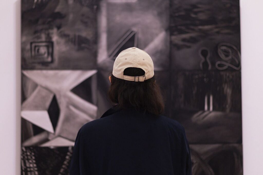 Una persona con una gorra de béisbol de color beige es vista desde atrás, mirando de cerca una gran obra de arte en blanco y negro colgada en una pared.