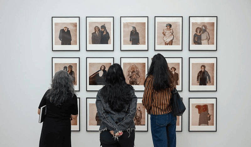 Three women stand with their backs to the camera, looking at rows of framed portraits of masked and costumed figures hanging on a white gallery wall.