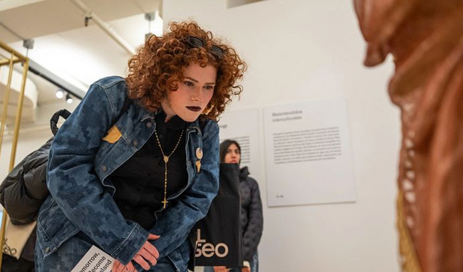 A woman with curly reddish-brown hair wearing a denim jacket leans forward to examine an artwork inside El Museo