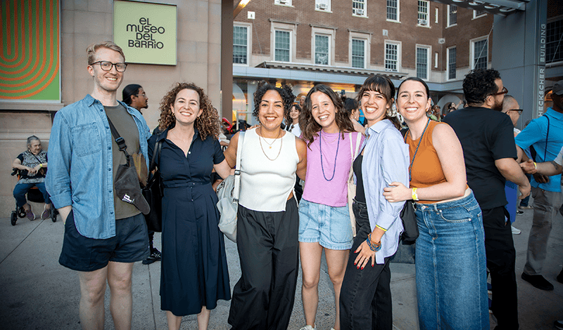 Un grupo de seis adultos jóvenes sonrientes posa junto al aire libre para una fotografía frente a El Museo del Barrio.