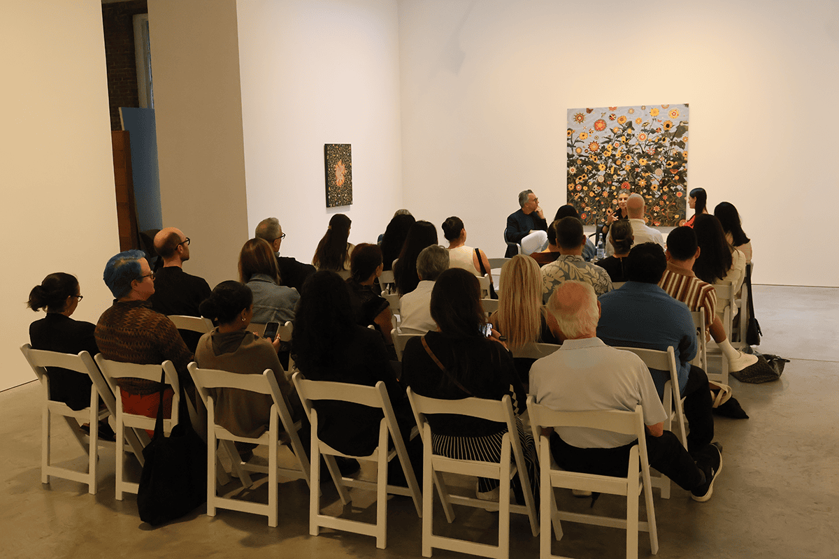 Un público se sienta en sillas plegables blancas en una galería de arte escuchando una presentación de panel frente a una gran pintura floral.