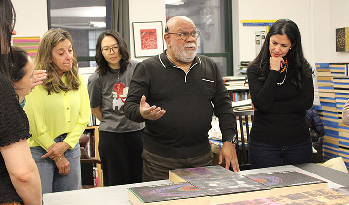 A man speaks and gestures while presenting artwork on a table to an attentive group of women gathered in a studio.
