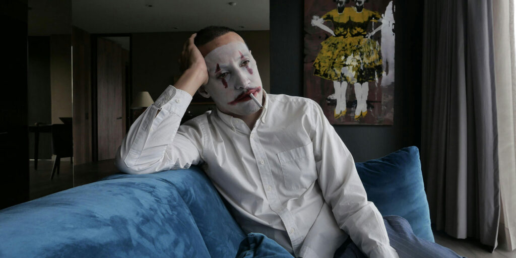 A man wearing clown makeup and a white shirt sits on a blue sofa smoking a cigarette