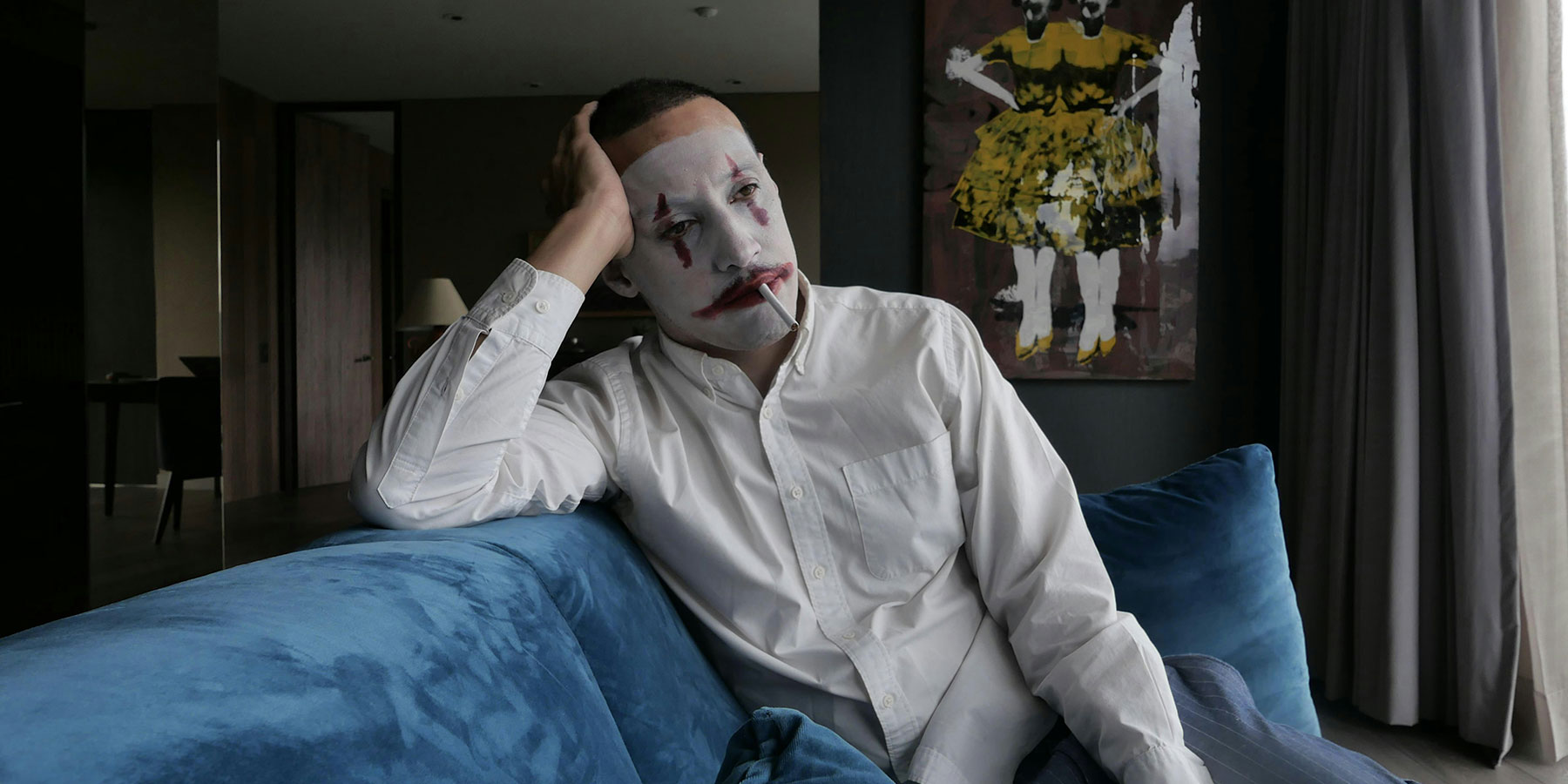 A man wearing clown makeup and a white shirt sits on a blue sofa smoking a cigarette