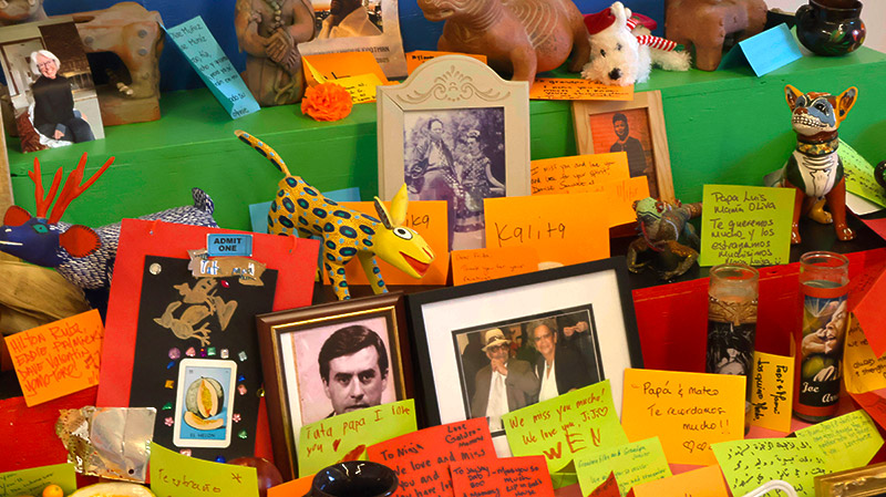 A colorful memorial altar displays framed photographs, animal figurines, candles, and handwritten notes on brightly colored paper.