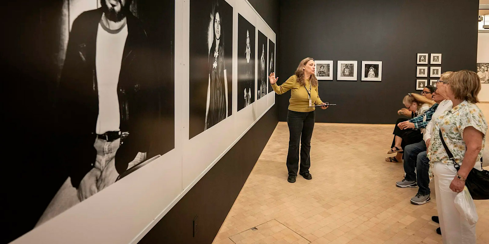 A museum tour guide gestures toward the artwork while explaining details to a group of attentive visitors.