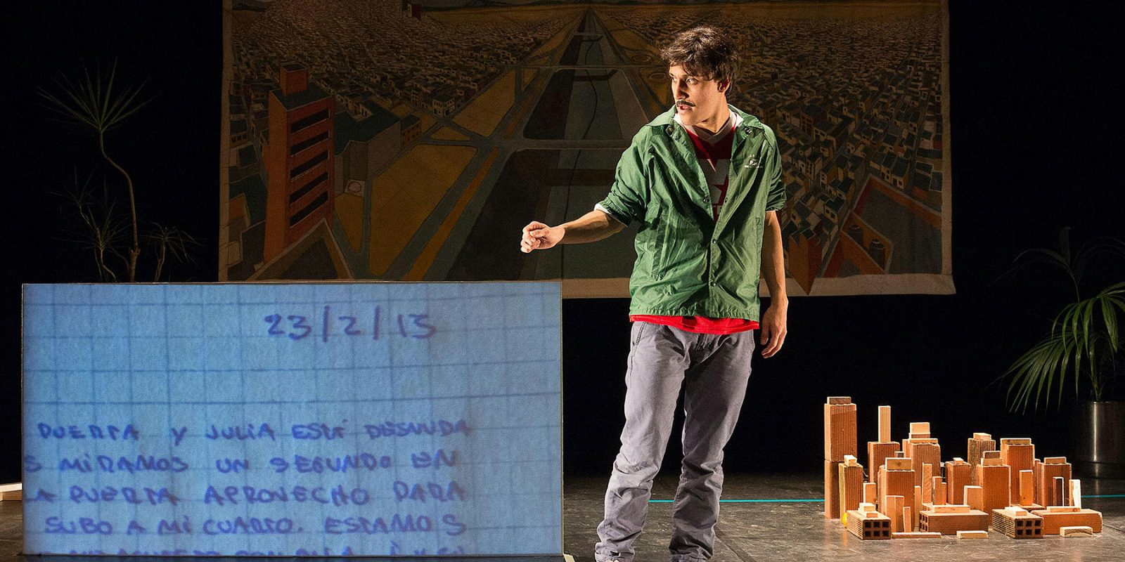 A man in a green jacket performs on a stage set with a large handwritten note and a miniature block city.