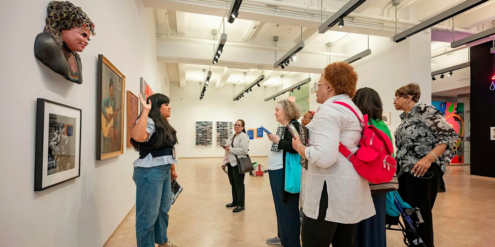 A museum tour guide gestures toward the artwork while explaining details to a group of attentive visitors.