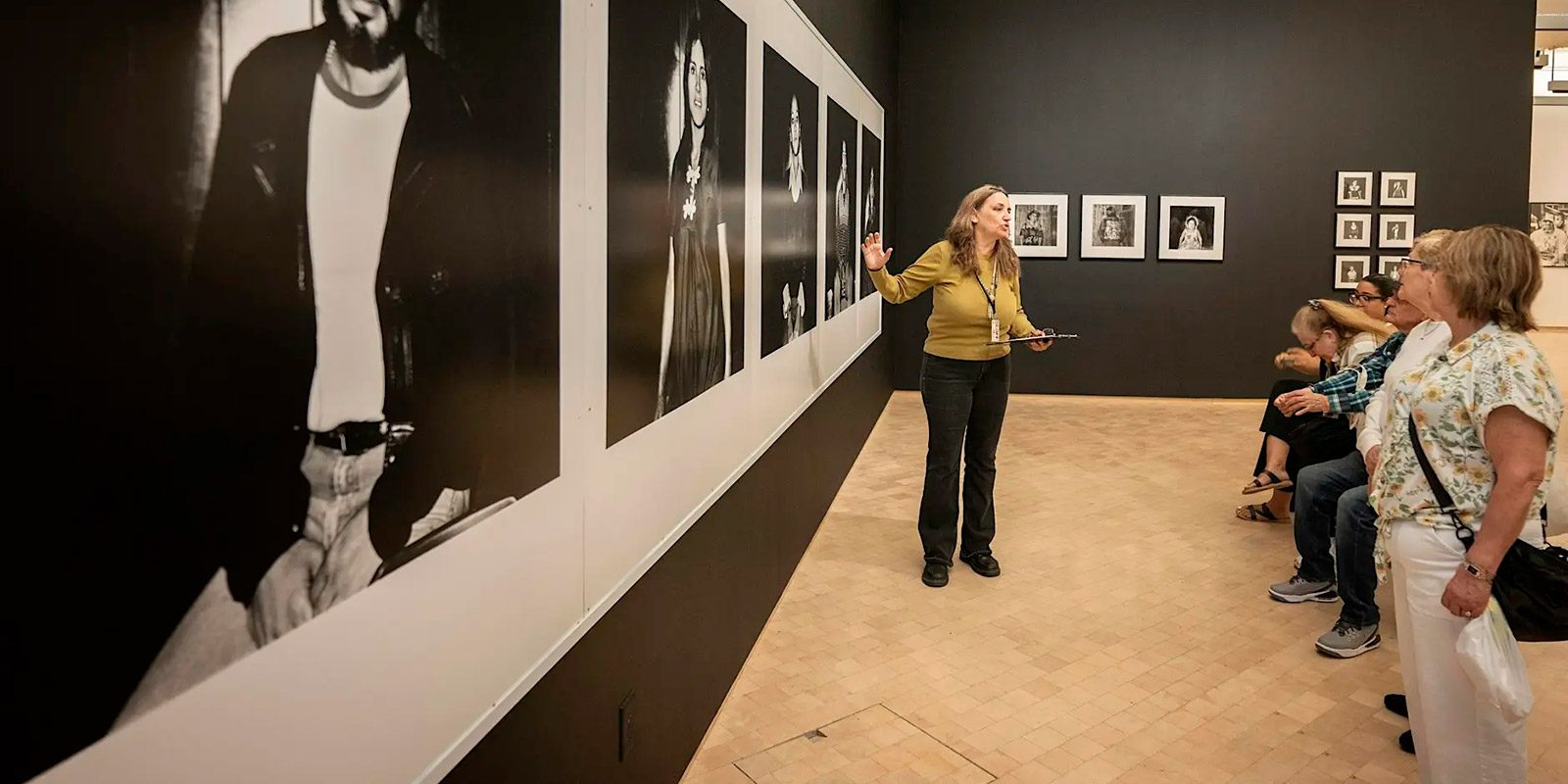A museum tour guide gestures toward the artwork while explaining details to a group of attentive visitors.