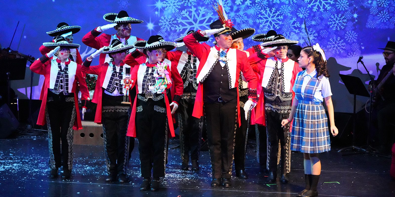 A group of performers in festive red-and-black mariachi-style outfits and sombreros salute on stage alongside a young girl in a blue plaid dress against a snowy backdrop.