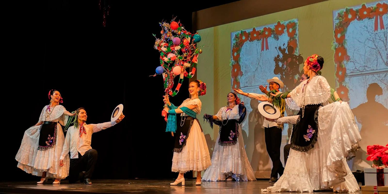 Dancers in traditional Mexican white dresses perform on stage, with one holding a decorated festive branch surrounded by balloons and ribbons.